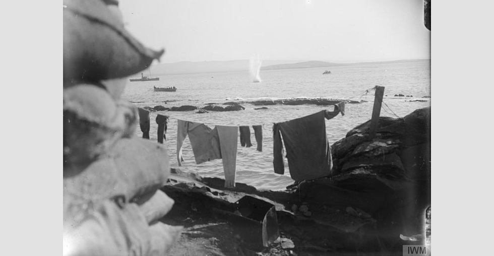 A shell from a Turkish battery plunging into the sea at the Dardanelles, after missing its target - the boat load of troops to the left. Note the laundry drying up in the foreground.